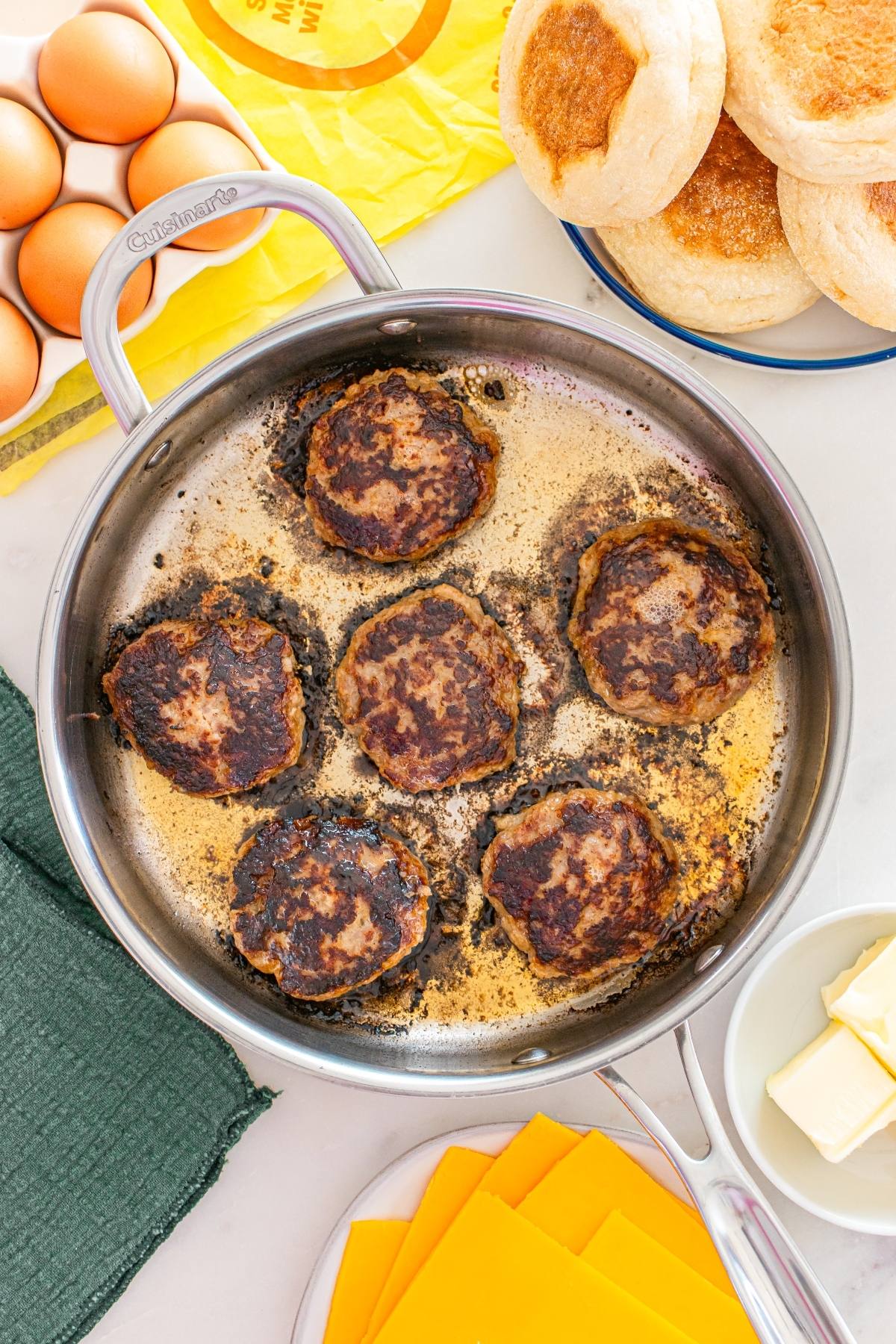 Six browned breakfast sausage patties cook in a stainless steel pan, surrounded by eggs, cheese, muffins, and butter.