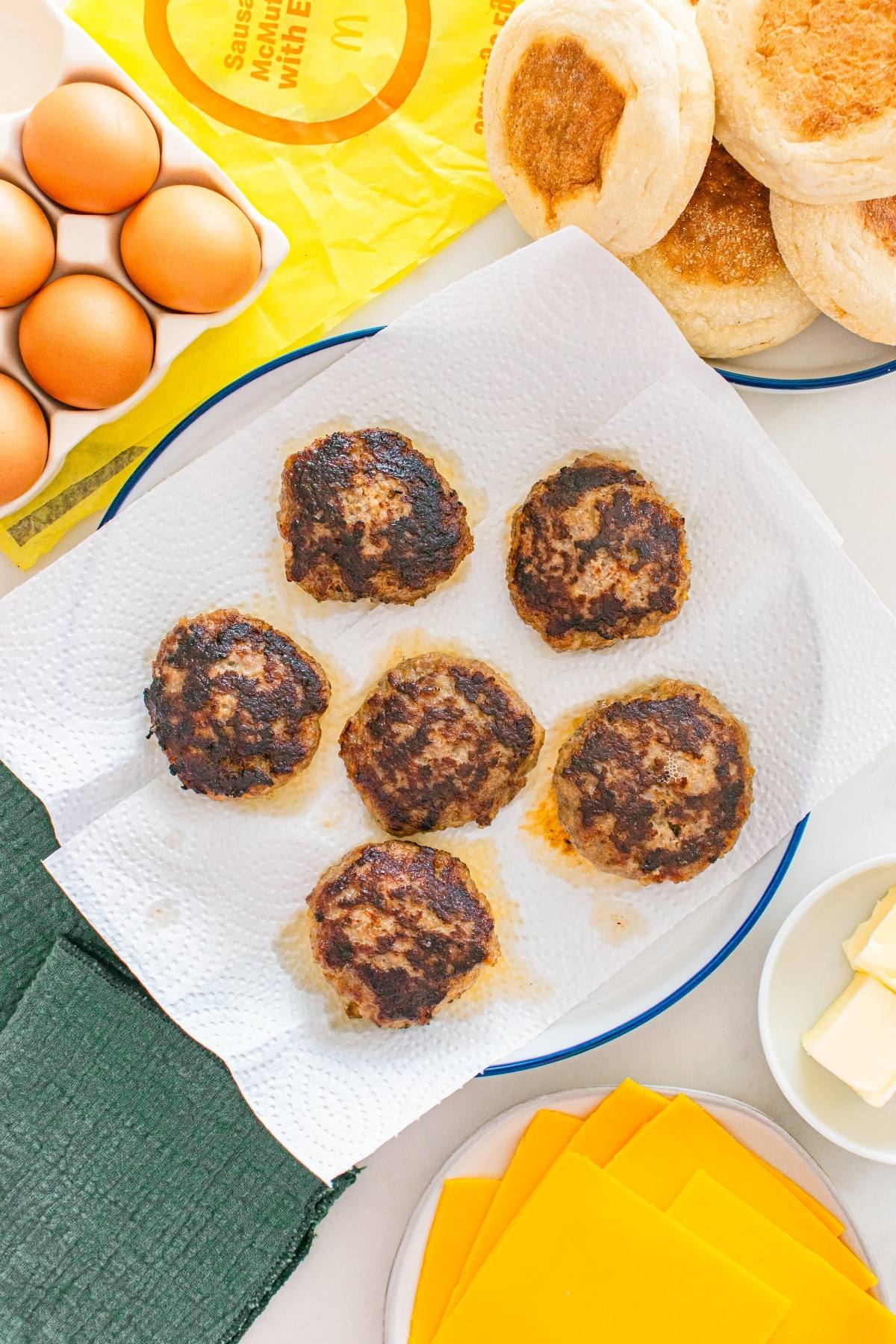 Six cooked sausage patties on paper towels, with eggs, english muffins, cheese, and butter nearby.