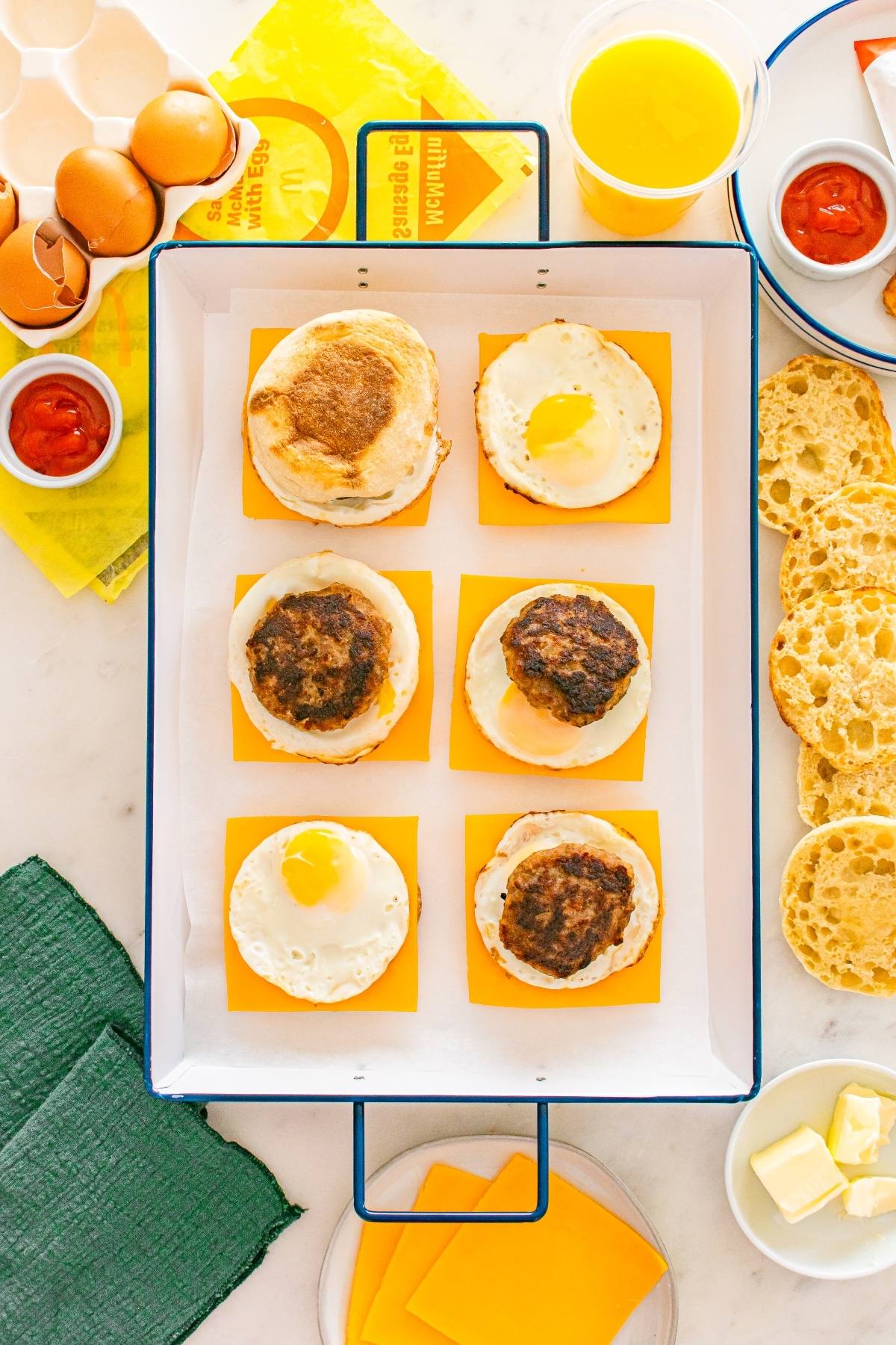 Six breakfast sandwiches with eggs, sausage, and cheese on english muffins, with orange juice and condiments nearby.