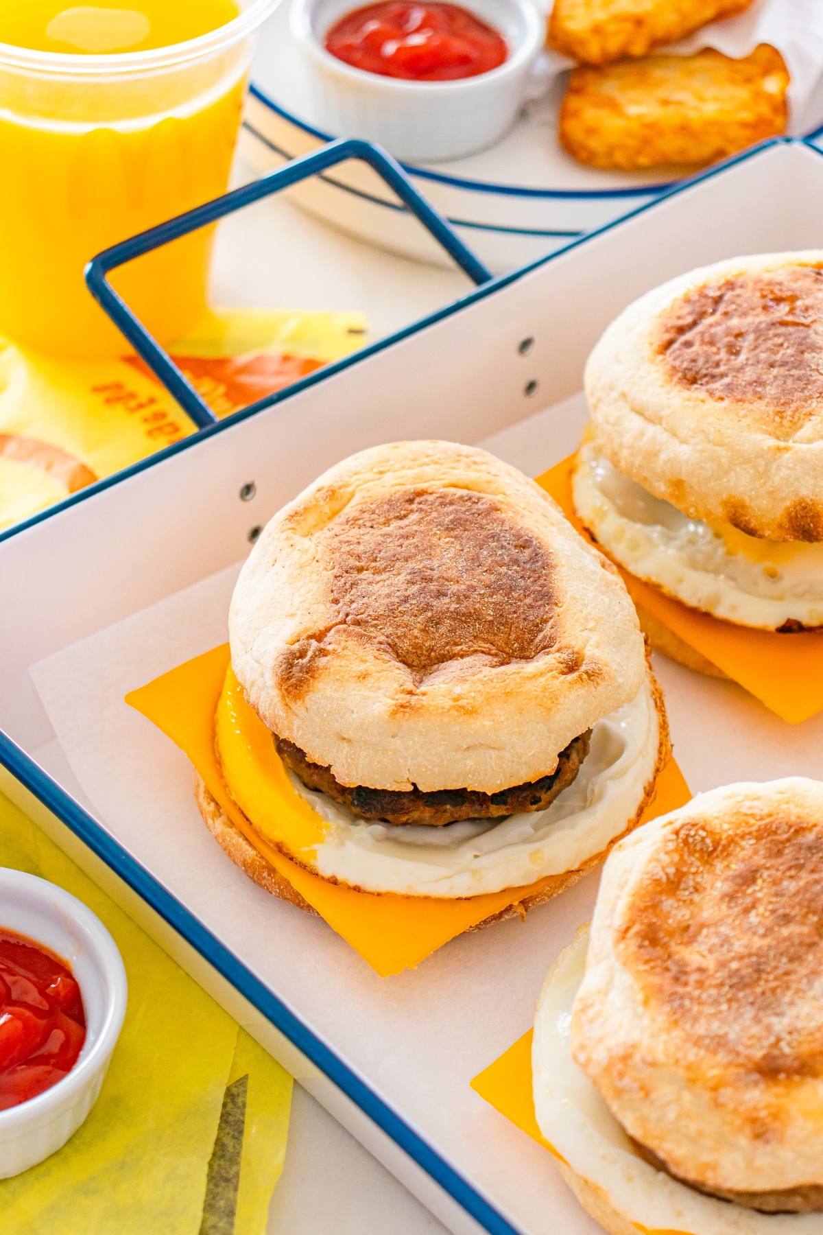 Breakfast sandwiches with egg, cheese, and sausage on english muffins, served with ketchup and orange juice.