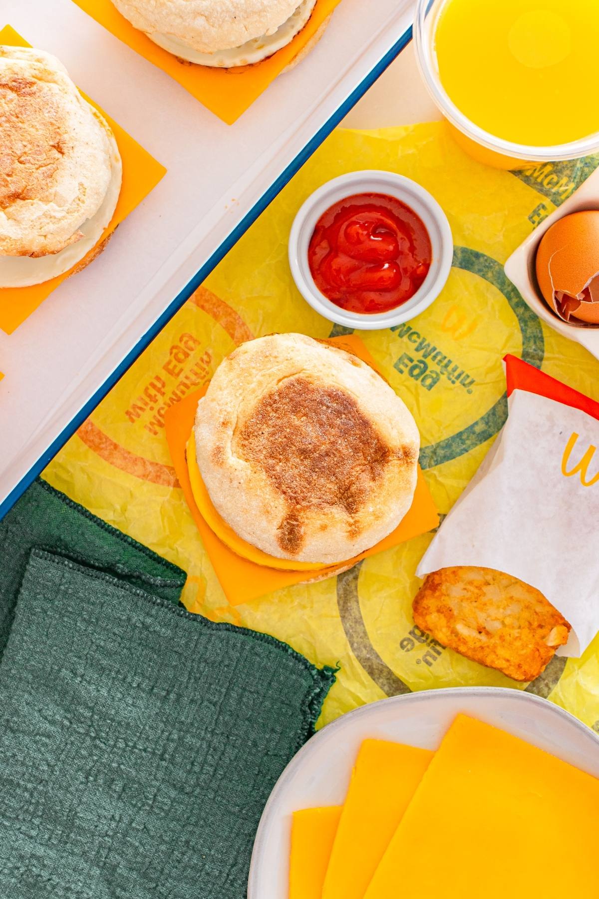 Breakfast with an english muffin sandwich, cheddar cheese slices, hash brown, ketchup, and a cup of orange juice.