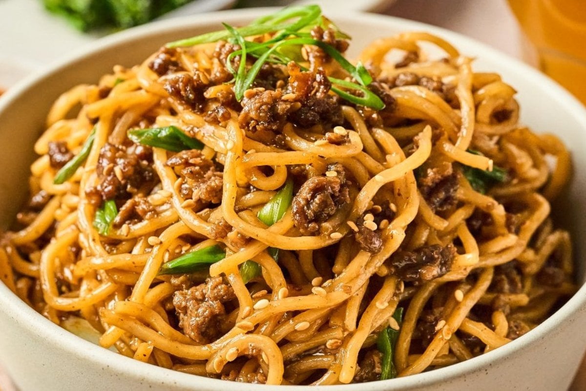 A bowl of noodles with ground meat, green onions, and sesame seeds, served in a light-colored bowl.