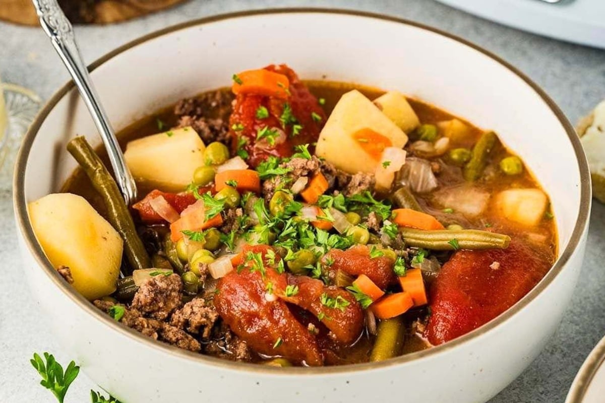 A bowl of vegetable beef soup with potatoes, carrots, green beans, peas, tomatoes, and fresh parsley on top.