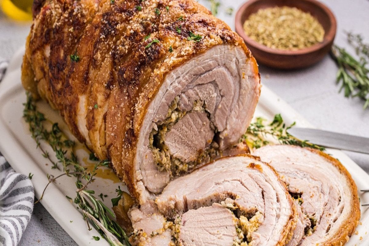 Sliced stuffed pork roast on a platter, garnished with herbs and served with seasoning in a small bowl.