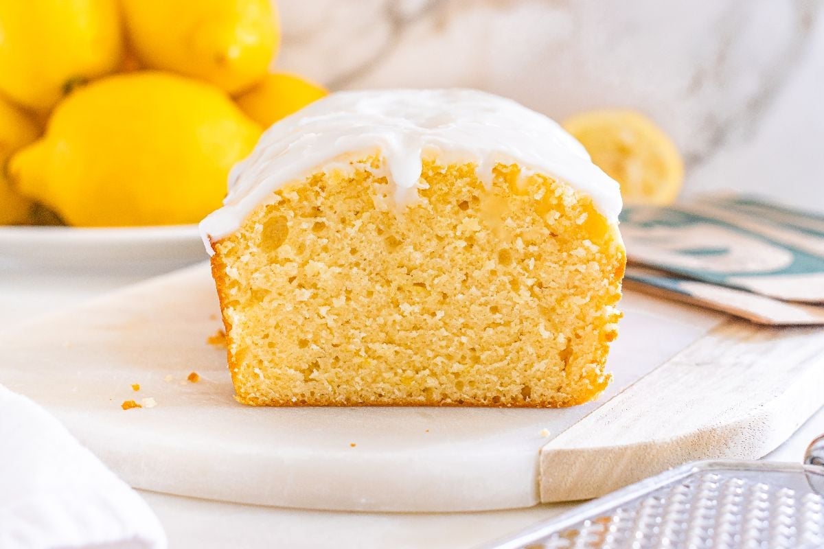 A sliced lemon loaf with white icing on top sits on a round white board, with lemons in the background.