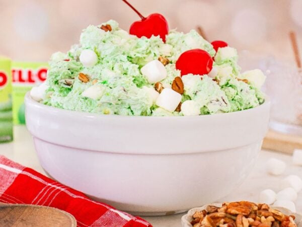 A white bowl of Watergate Salad—green pistachio fluff with marshmallows, nuts, and cherries on top—sits next to Jell-O boxes.