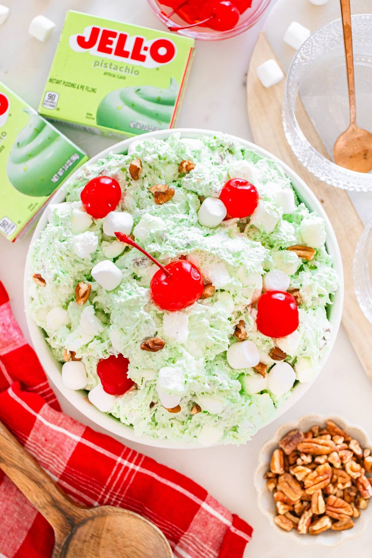 A bowl of Watergate salad topped with cherries, marshmallows, and chopped pecans, with Jell-O box nearby.