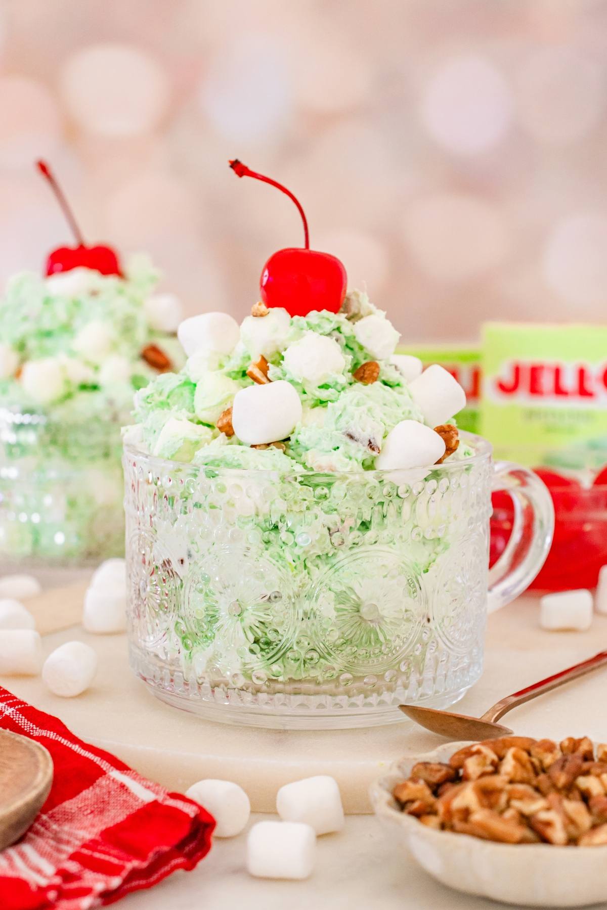 A glass cup filled with green Watergate salad, mini marshmallows, nuts, and topped with a cherry.