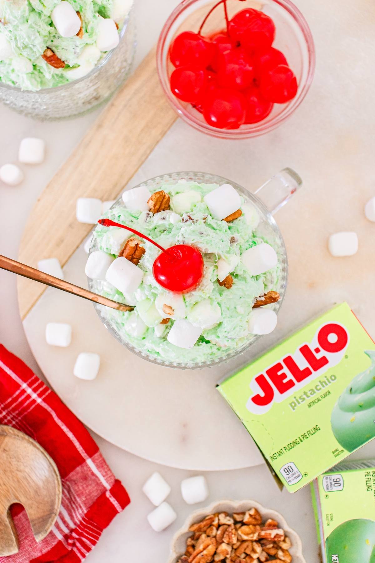 A bowl of Watergate salad with marshmallows, pecans, and a cherry, next to Jell-O mix and cherries.