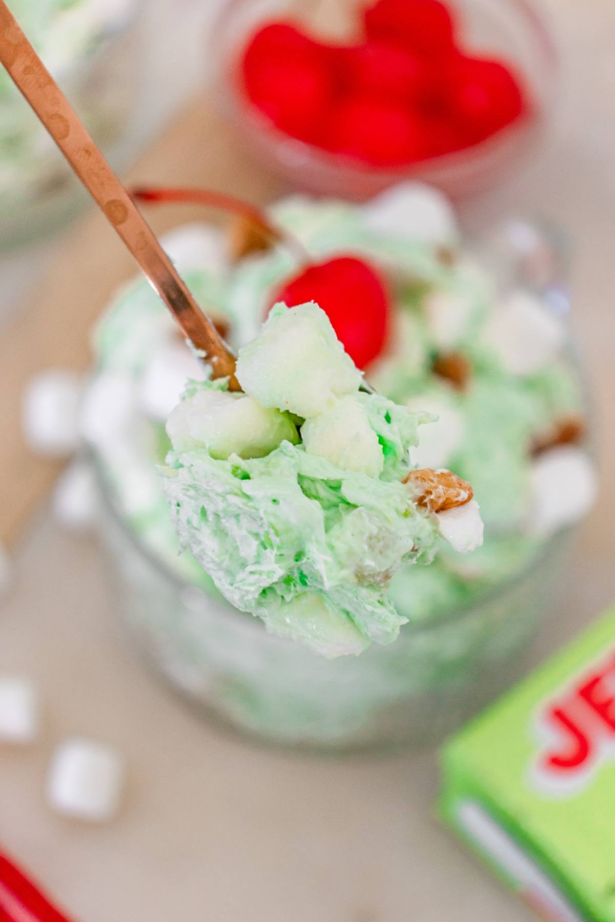 A spoonful of green Watergate salad with marshmallows and a cherry, held above a glass bowl.