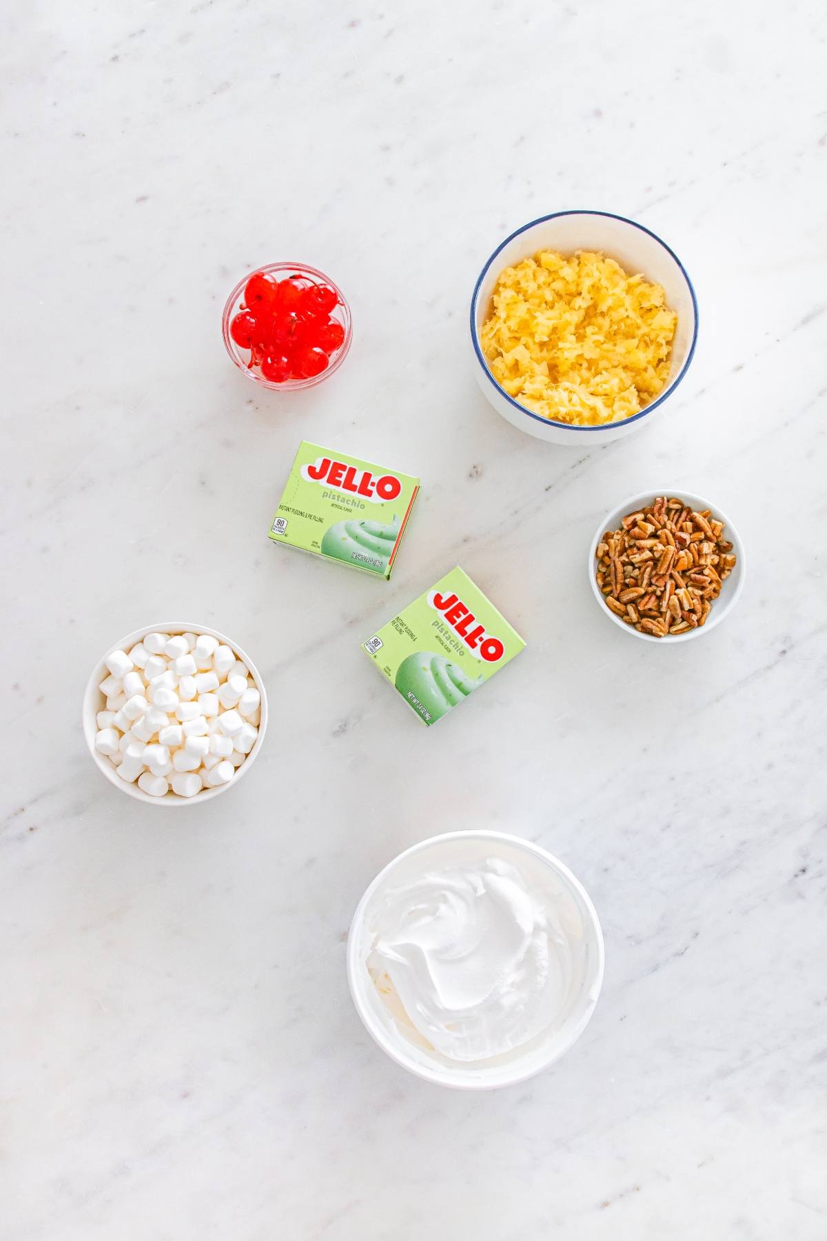 Seven bowls with ingredients: cherries, pineapple, pistachio pudding, pecans, marshmallows, and whipped topping.
