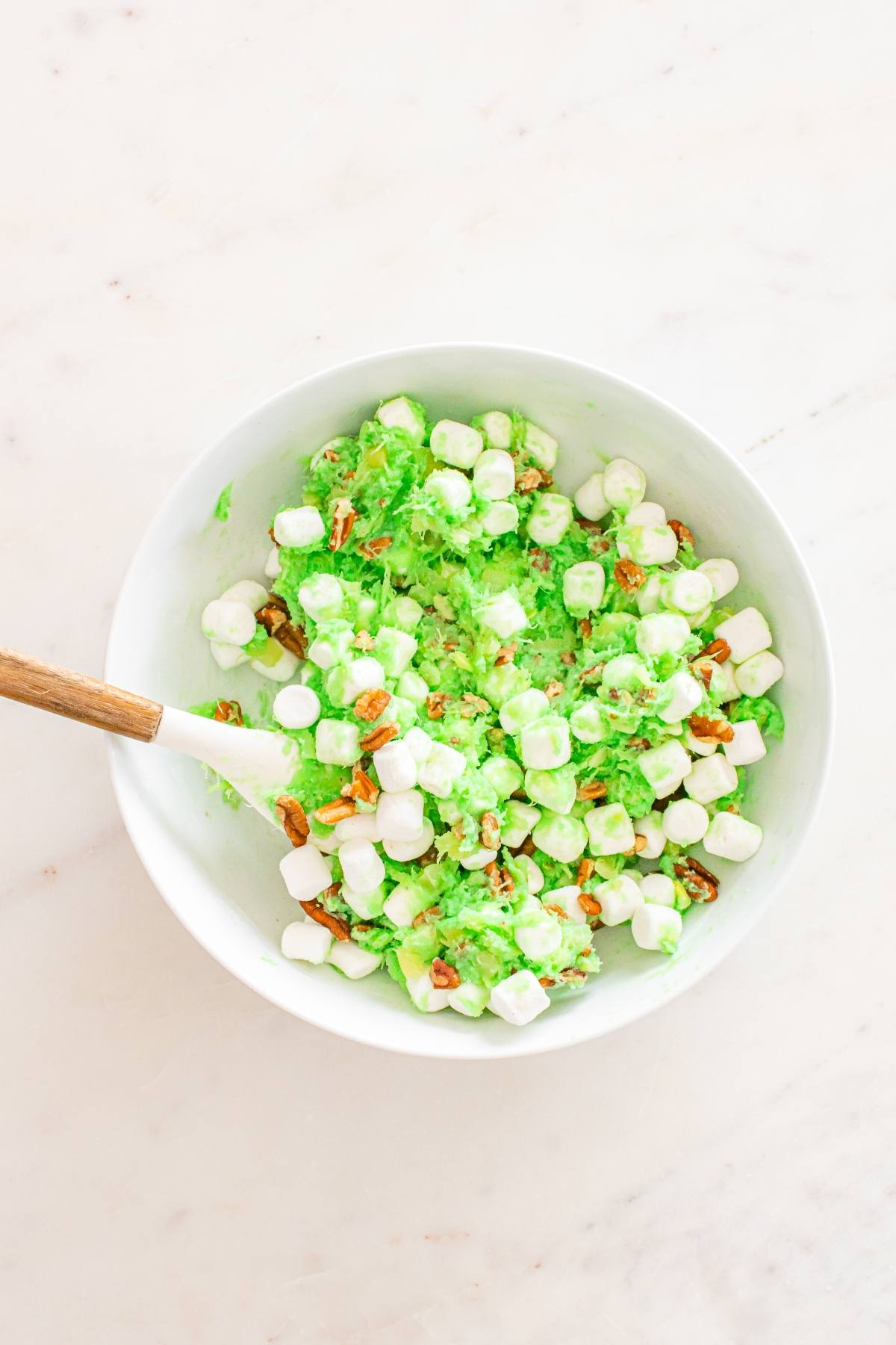 Bowl of green ambrosia salad with mini marshmallows, chopped pecans, and a white spoon on a marble surface.