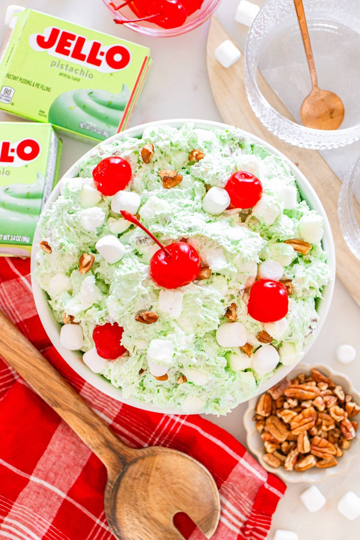 Bowl of Watergate salad topped with cherries, marshmallows, and pecans, with Jello boxes and ingredients nearby.