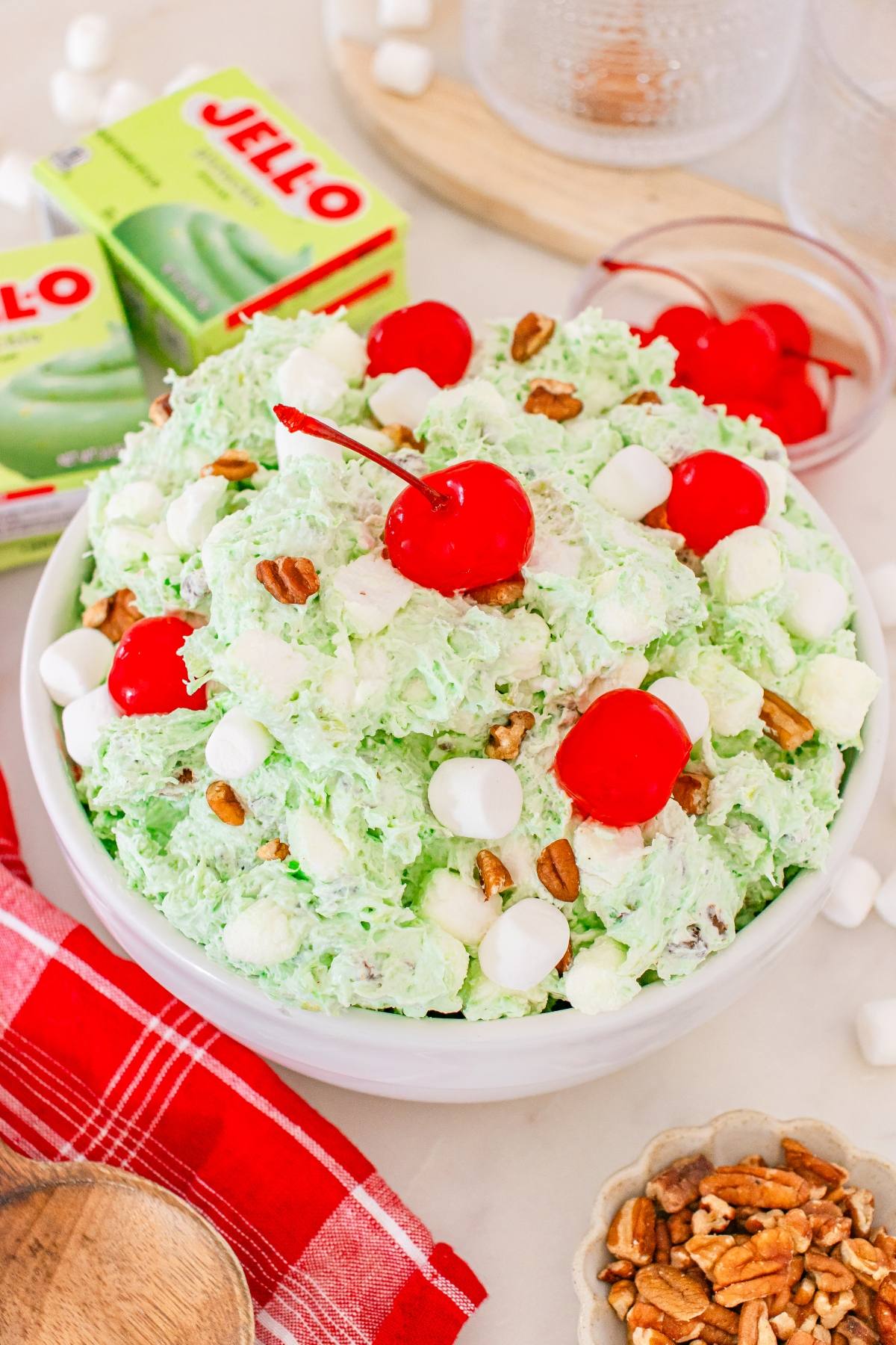 A bowl of Watergate salad with marshmallows, pecans, and cherries, surrounded by Jell-O boxes and ingredients.