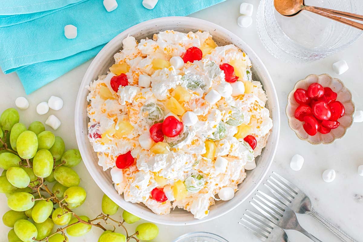 A bowl of classic ambrosia salad with marshmallows, cherries, pineapple, and grapes sits on a white table.