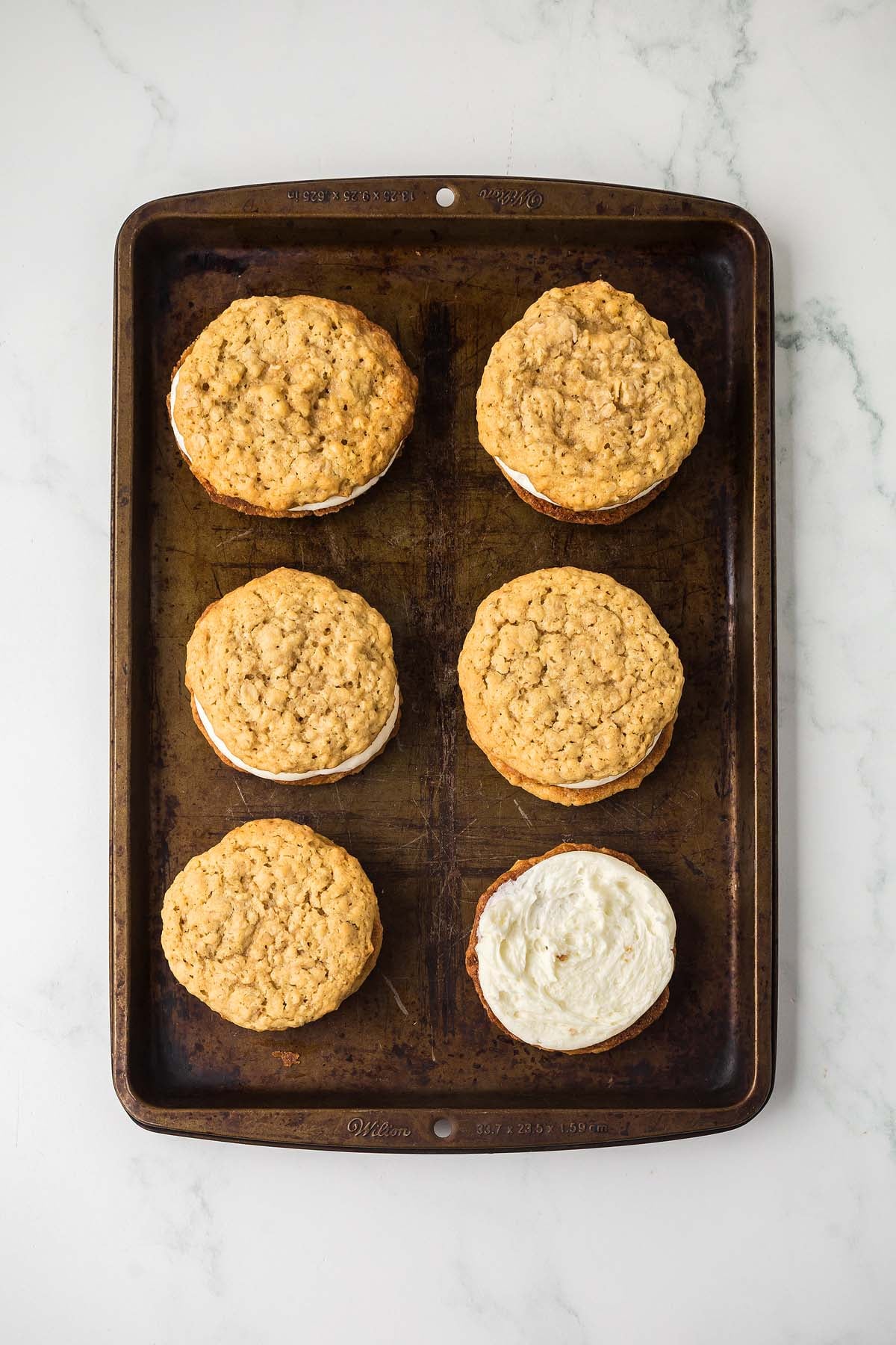 Six Homemade Oatmeal Cream Pies rest on a baking sheet, one with its creamy filling exposed, inviting you to indulge, all set atop a marble countertop.