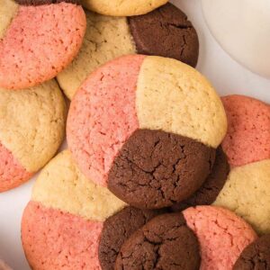 Assorted Neapolitan Cookies in pink, brown, and beige are displayed on a white surface next to a jar and scattered chocolate chips.