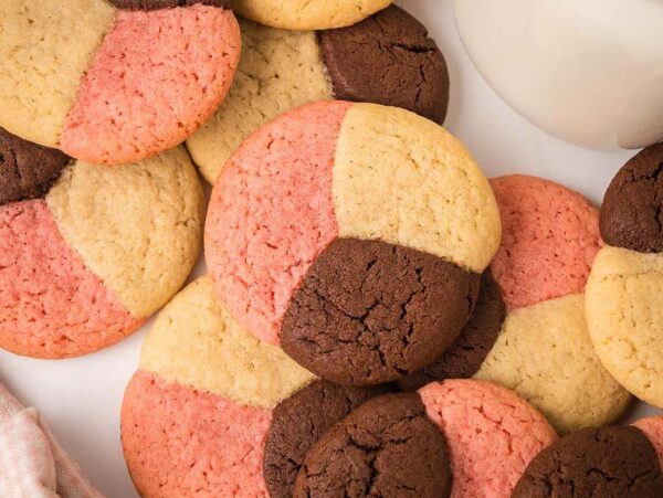 Assorted Neapolitan Cookies in pink, brown, and beige are displayed on a white surface next to a jar and scattered chocolate chips.