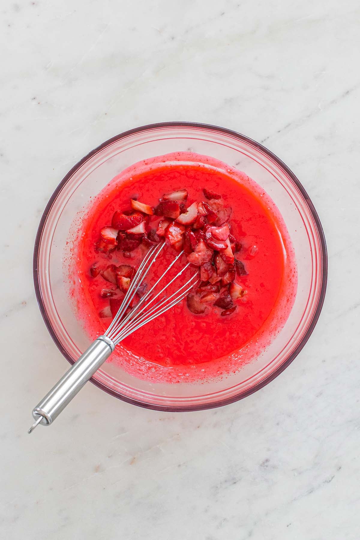 A glass bowl with chopped strawberries in red liquid, perfect for making strawberry chiffon squares, and a metal whisk on a white marble surface.
