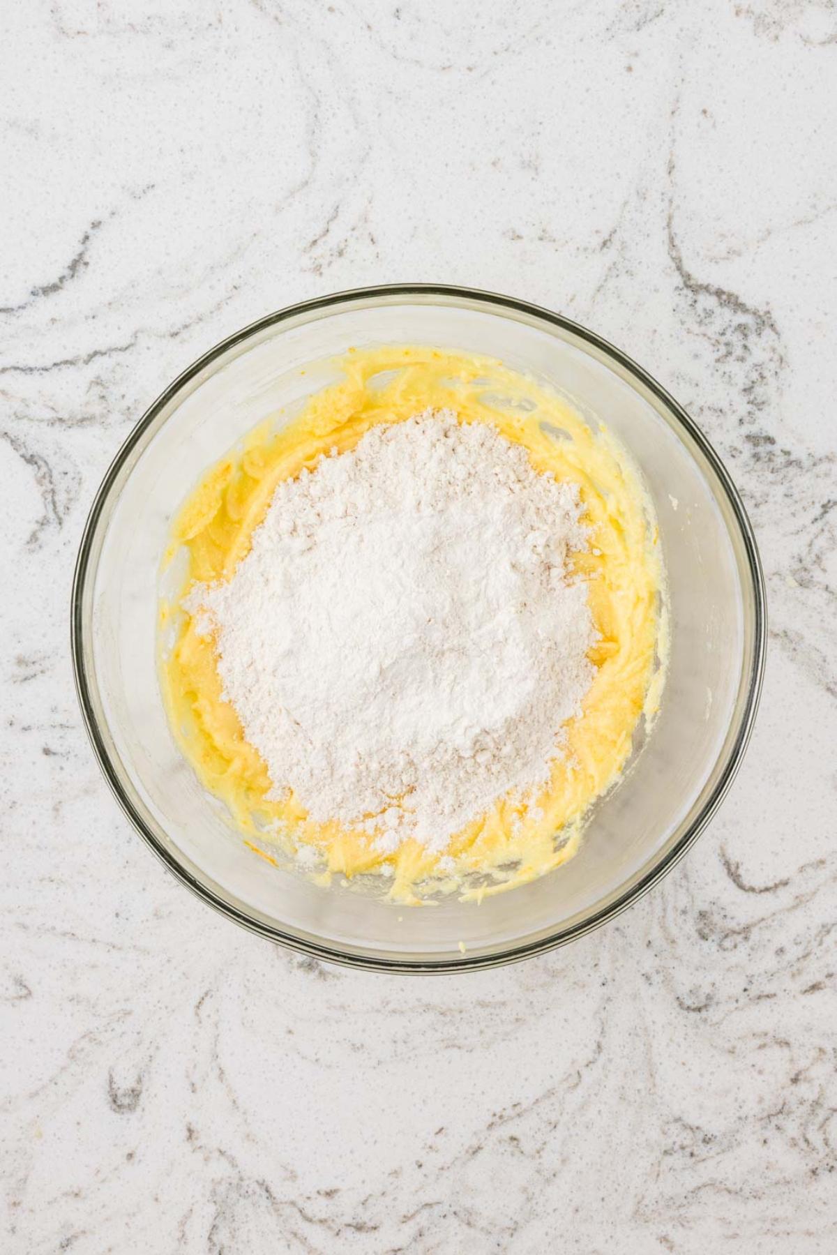 A glass bowl with creamed butter and sugar, topped with flour, on a white marble surface.