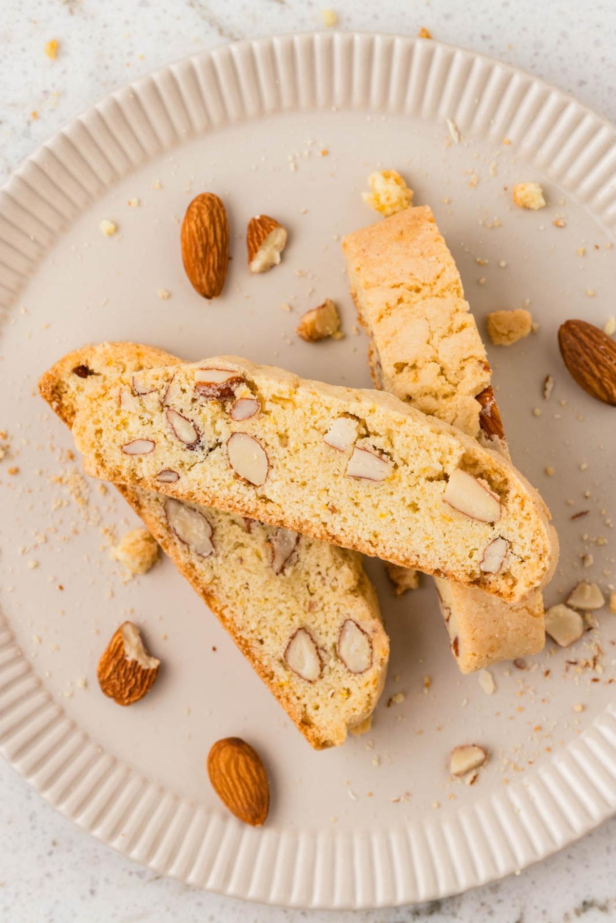 Two almond biscotti on a plate with whole almonds and crumbs scattered around.