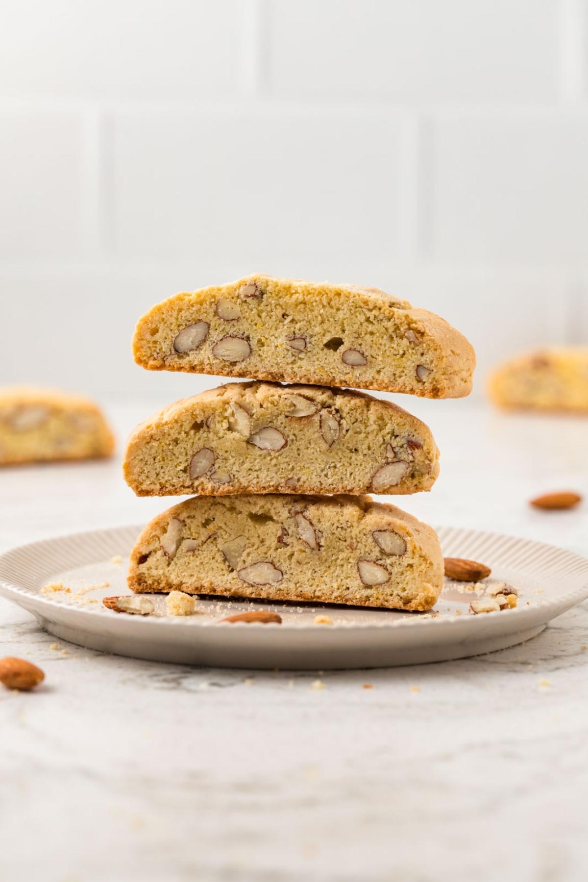 Three almond biscotti stacked on a plate, with more cookies and almonds in the background.
