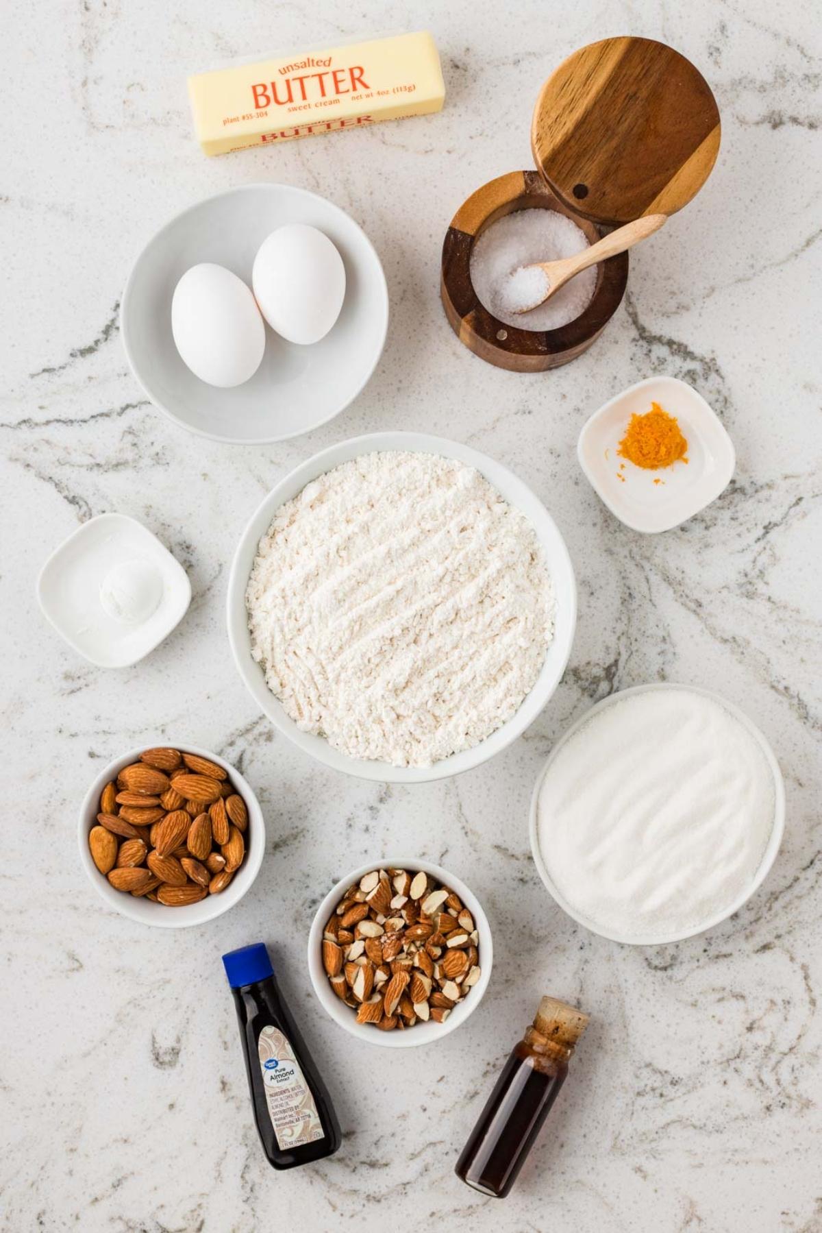 Baking ingredients on a marble counter: flour, eggs, butter, sugar, almonds, vanilla, orange zest, salt, and baking powder.