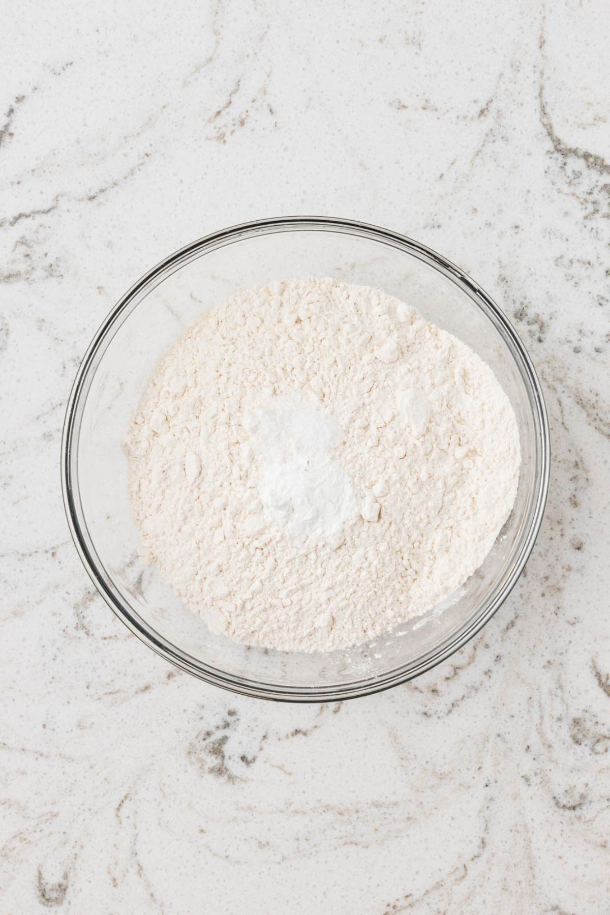 A glass bowl filled with flour and baking powder on a white marble surface.