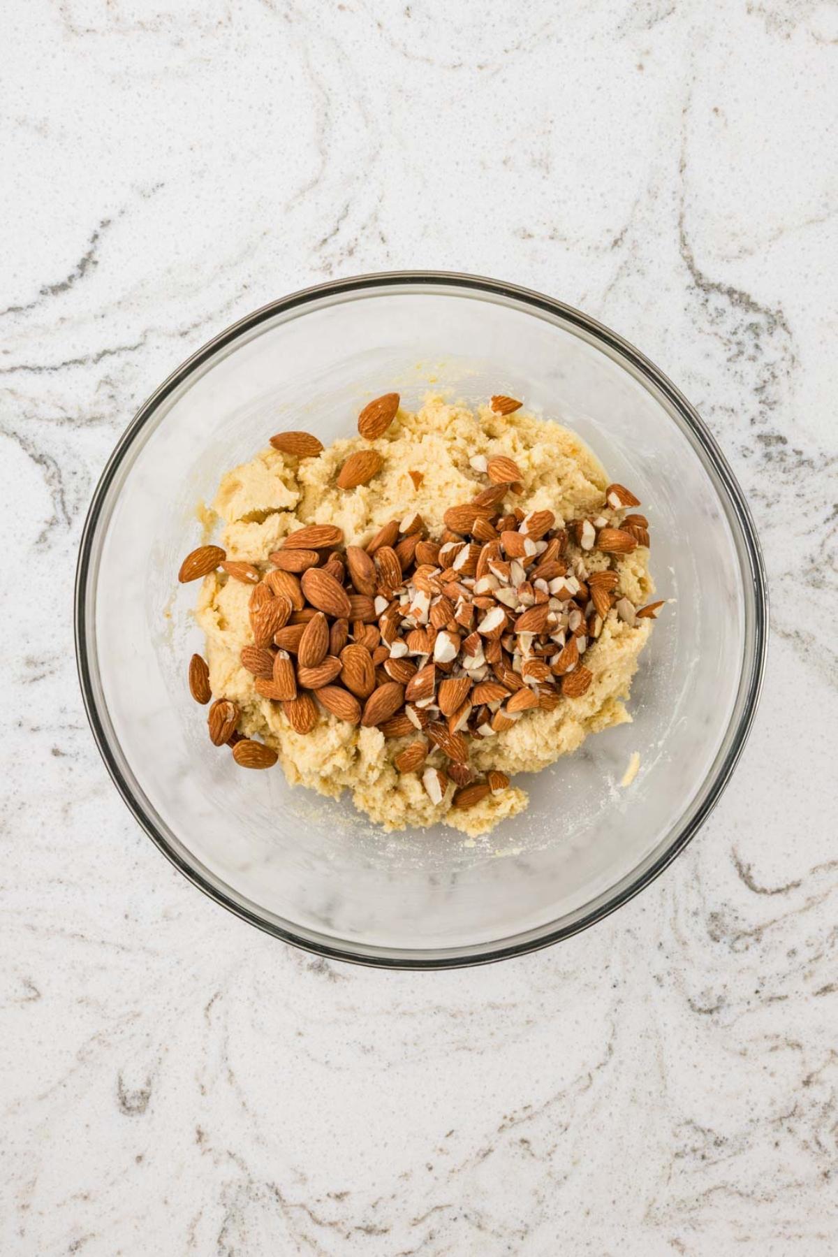 A glass bowl with cookie dough and chopped almonds on a white marble surface.