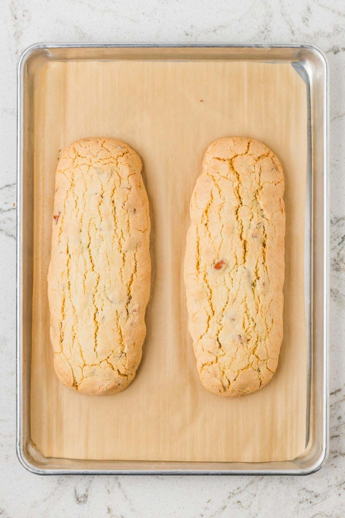 Two golden, cracked loaves of biscotti dough on a parchment-lined baking sheet.