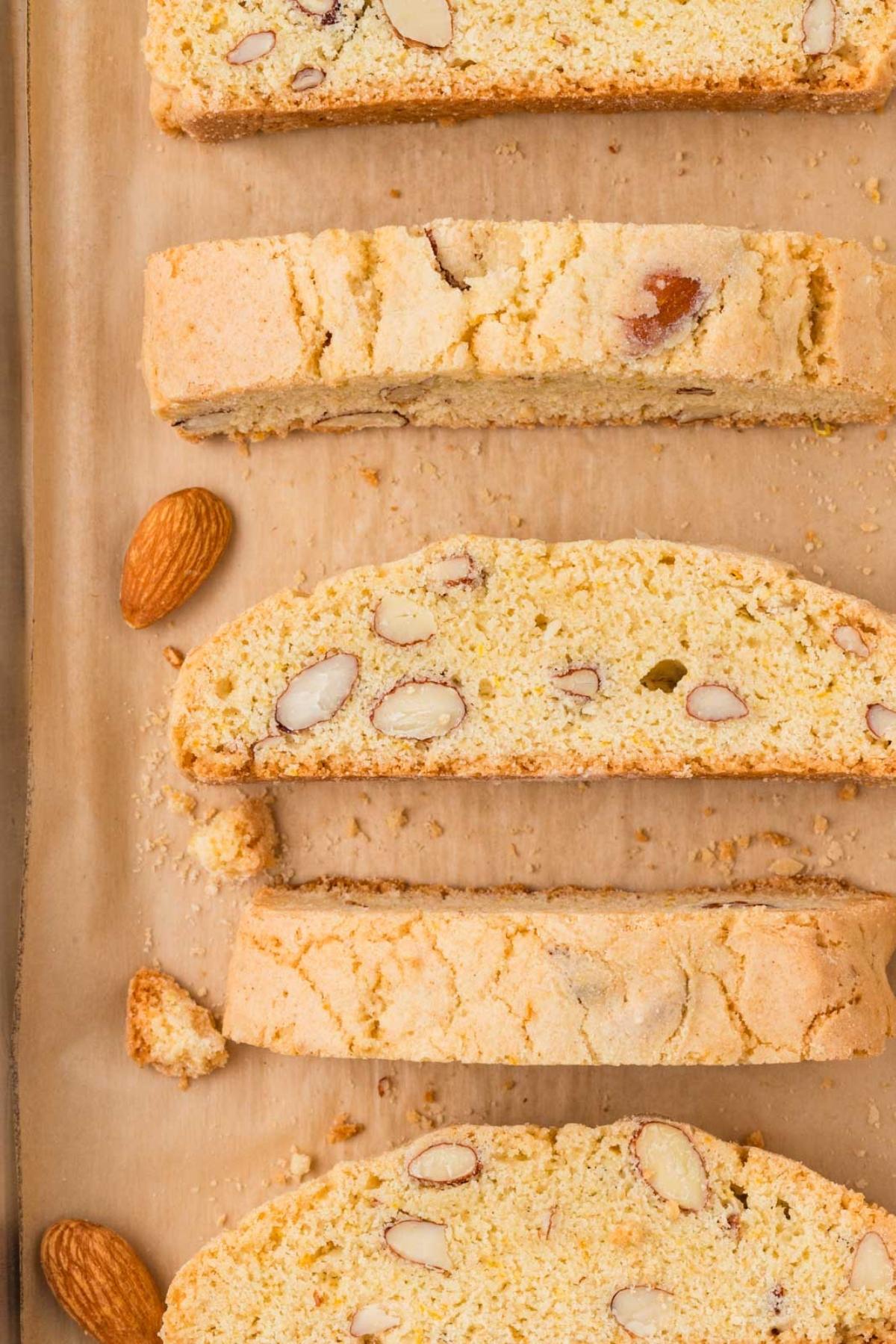Four slices of almond biscotti and whole almonds on brown parchment paper, viewed from above.