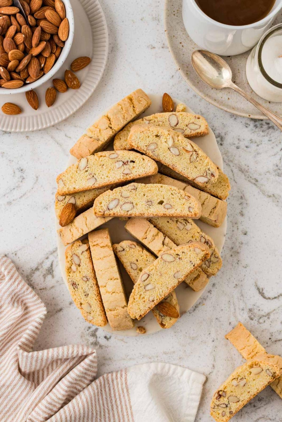 A plate of almond biscotti on a marble surface, with a bowl of almonds, coffee, and a striped napkin nearby.