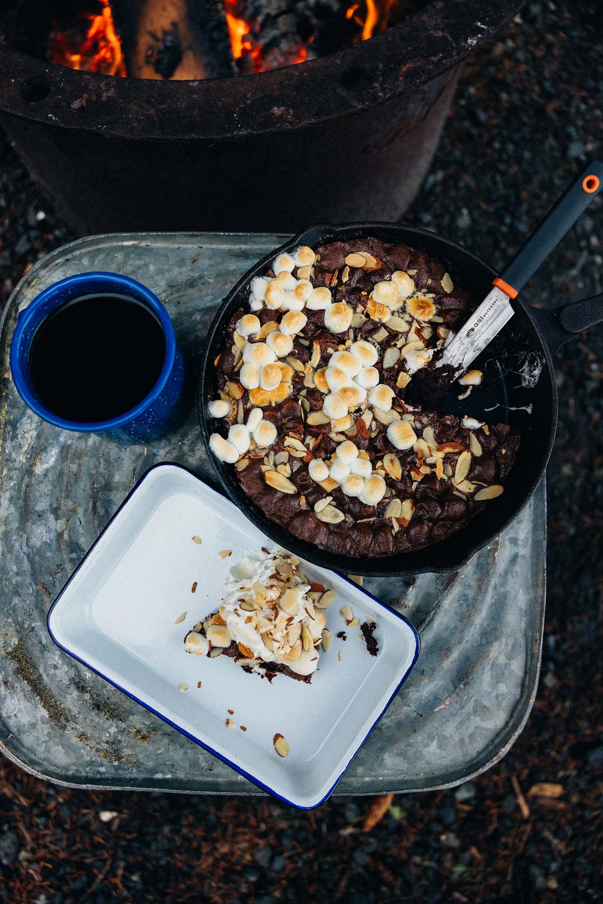 A campfire treat: cast iron caramel brownies studded with marshmallows and nuts, served next to coffee on a metal tray by the fire.