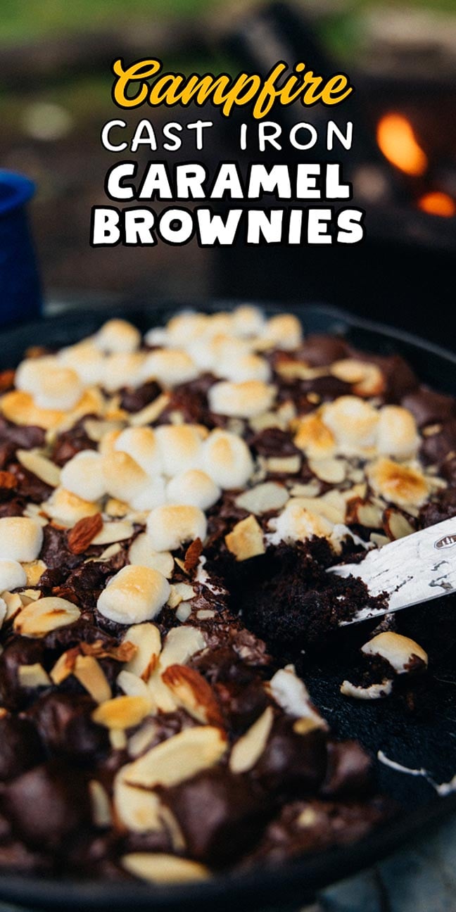 A skillet of Campfire Cast Iron Caramel Brownies topped with marshmallows and nuts sits beside the fire, ready to serve with a spoon.
