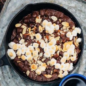 A Campfire Cast Iron Brownie, topped with toasted marshmallows and almond slices, sits on a metal surface next to a blue mug.