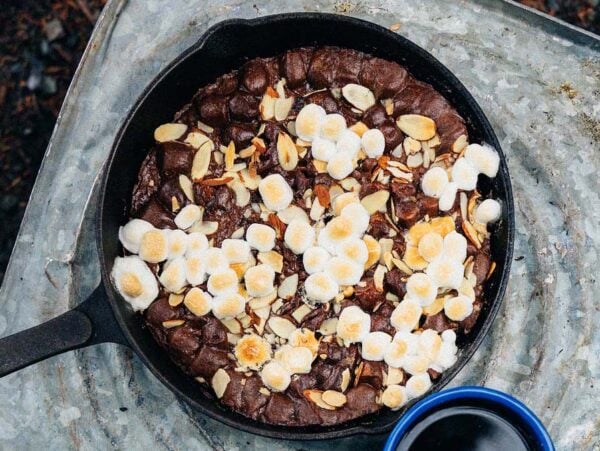 A Campfire Cast Iron Brownie, topped with toasted marshmallows and almond slices, sits on a metal surface next to a blue mug.