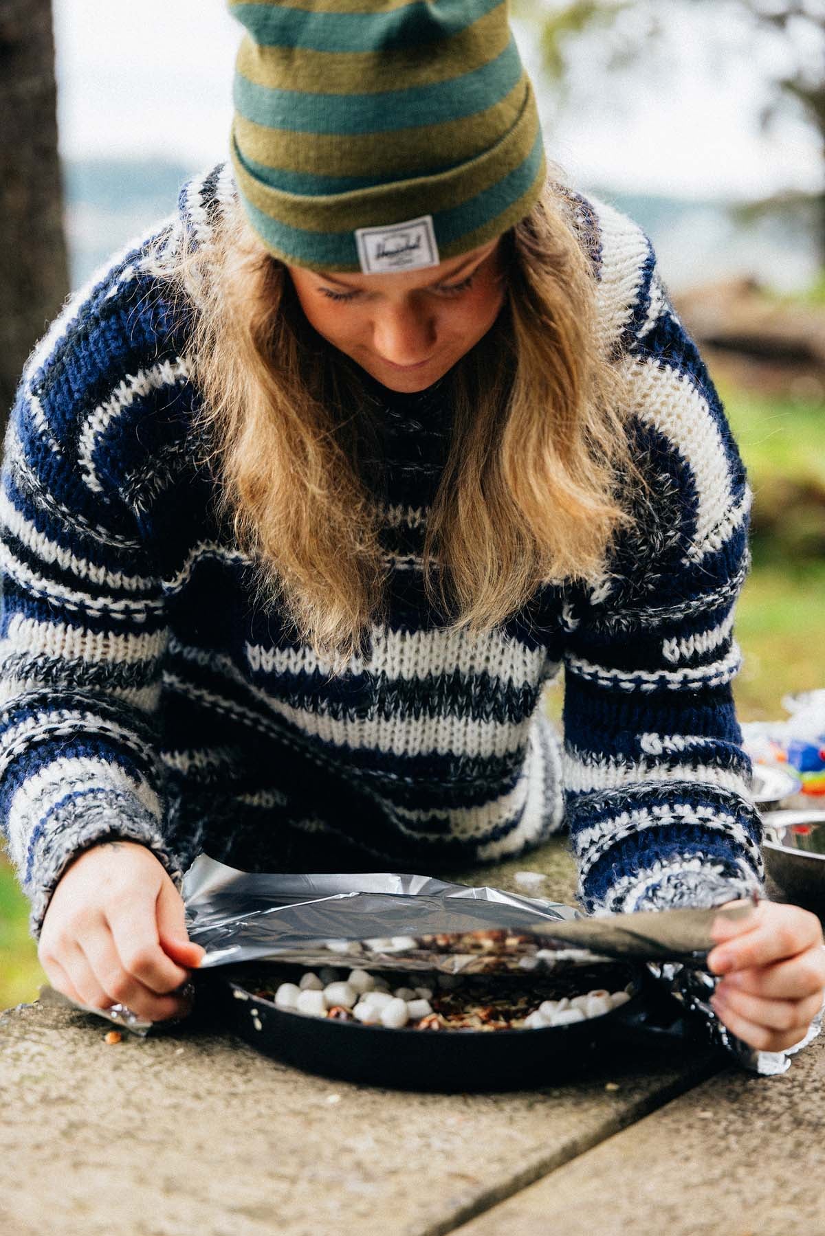 Person in a striped sweater and beanie lifts foil from a skillet with gooey marshmallows outdoors, preparing Campfire Cast Iron Caramel Brownies.