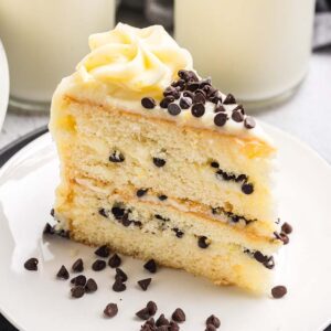 A slice of Cannoli Cake with chocolate chips and white frosting sits on a plate, alongside a glass of milk and extra chips for garnish.