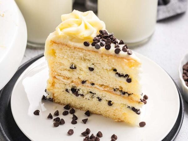 A slice of Cannoli Cake with chocolate chips and white frosting sits on a plate, alongside a glass of milk and extra chips for garnish.