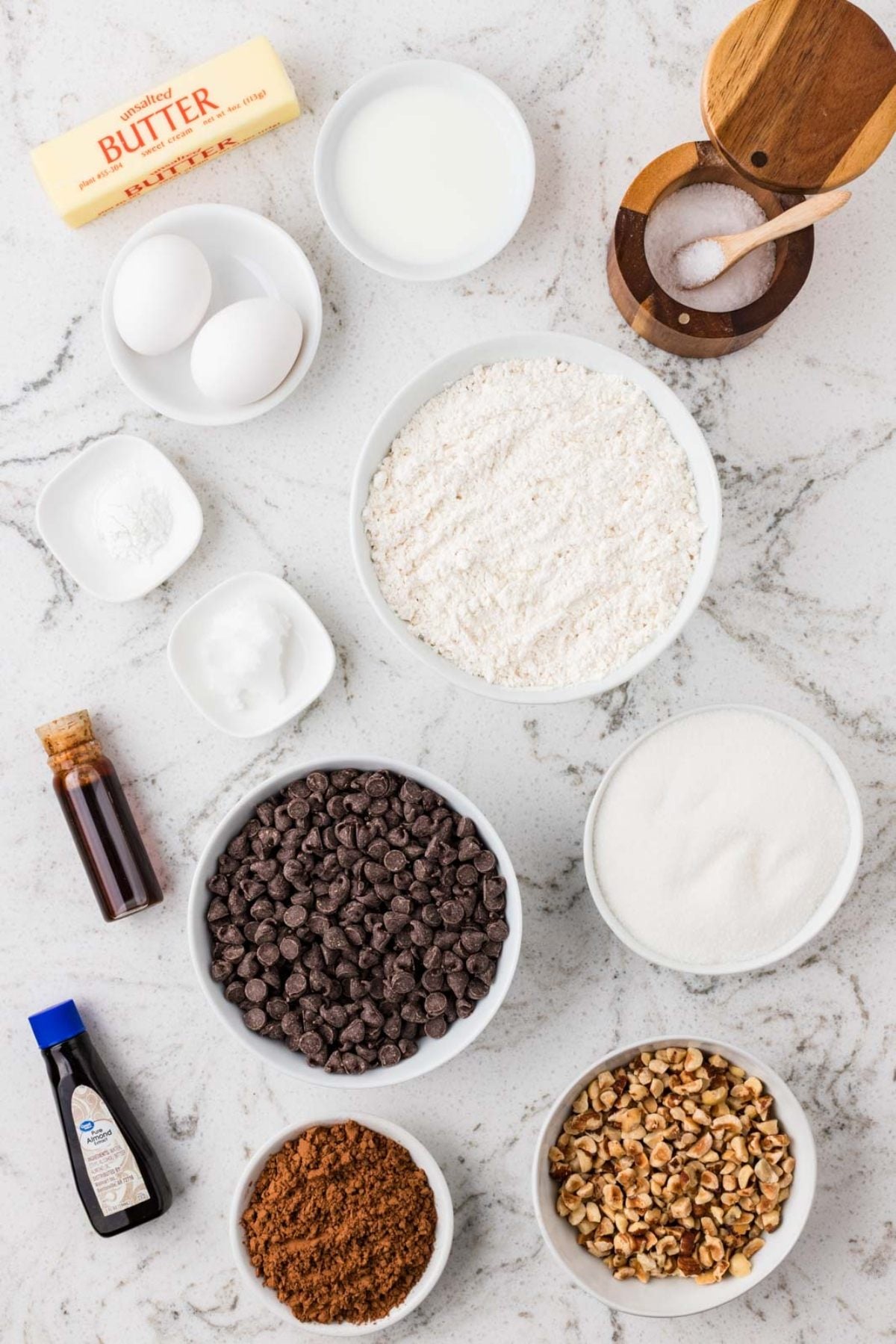 Bowls of chocolate chips, flour, sugar, nuts, eggs, butter, and other baking ingredients on a marble counter.