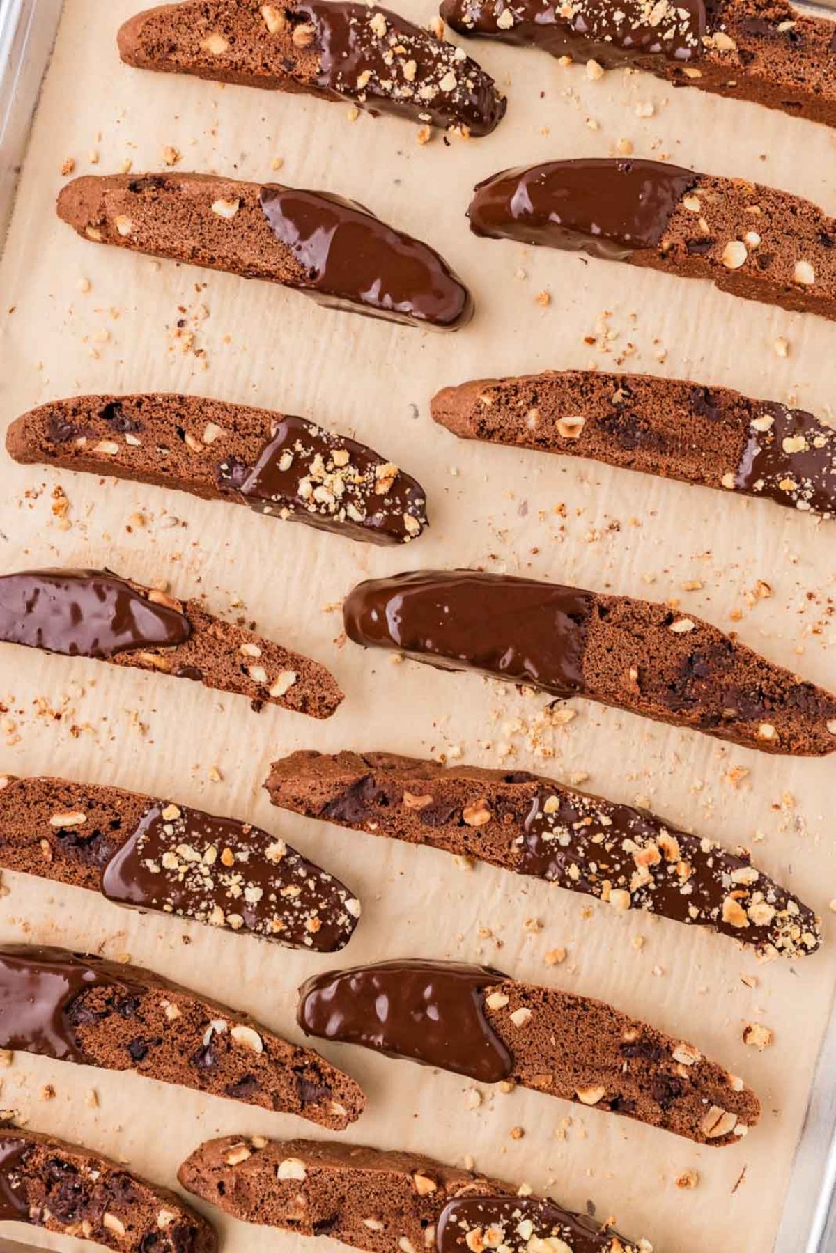Rows of biscotti cookies half-dipped in chocolate and sprinkled with nuts on parchment paper.