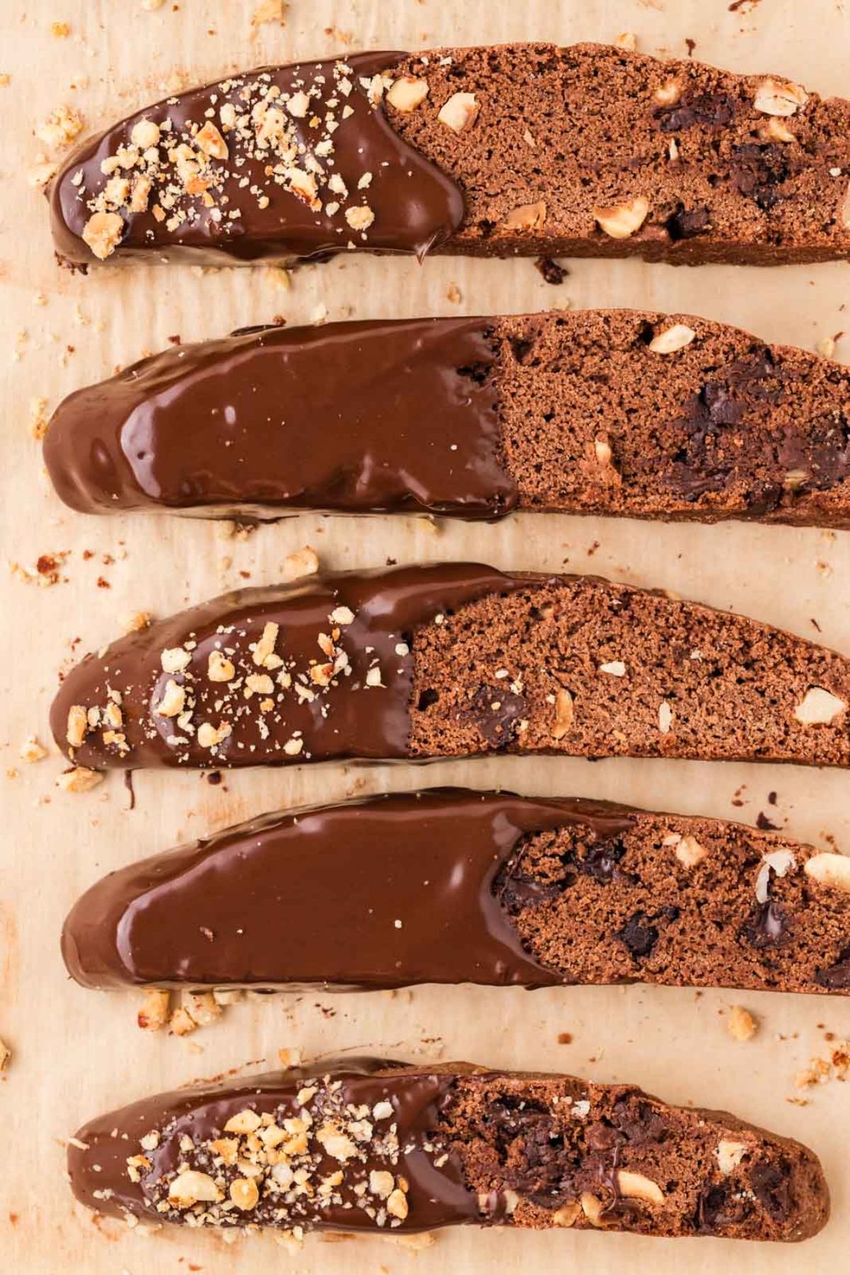 Five chocolate-dipped biscotti with chopped nuts on parchment paper, arranged in a row.