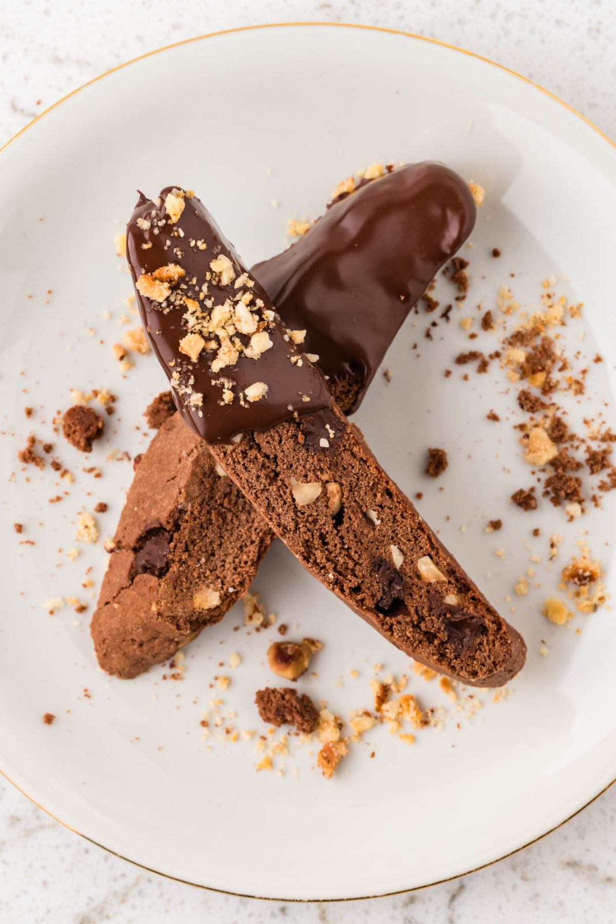 Two chocolate biscotti, one half-dipped in chocolate and sprinkled with nuts, on a white plate with crumbs.