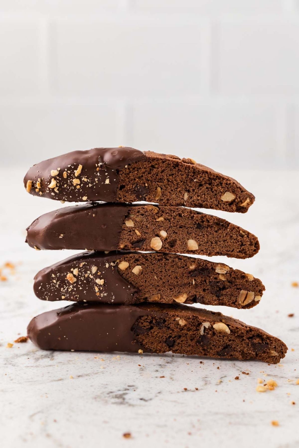 Four chocolate-dipped biscotti with nuts stacked on a light surface against a white background.