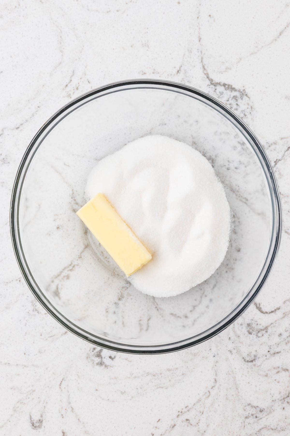 A glass bowl with a stick of butter and a pile of granulated sugar on a white marbled surface.