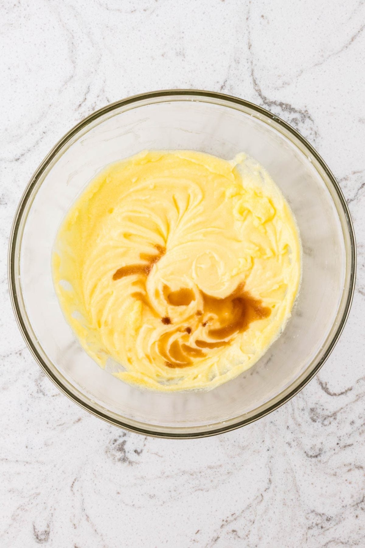 A glass bowl with yellow cake batter and a swirl of vanilla extract on a marble surface.