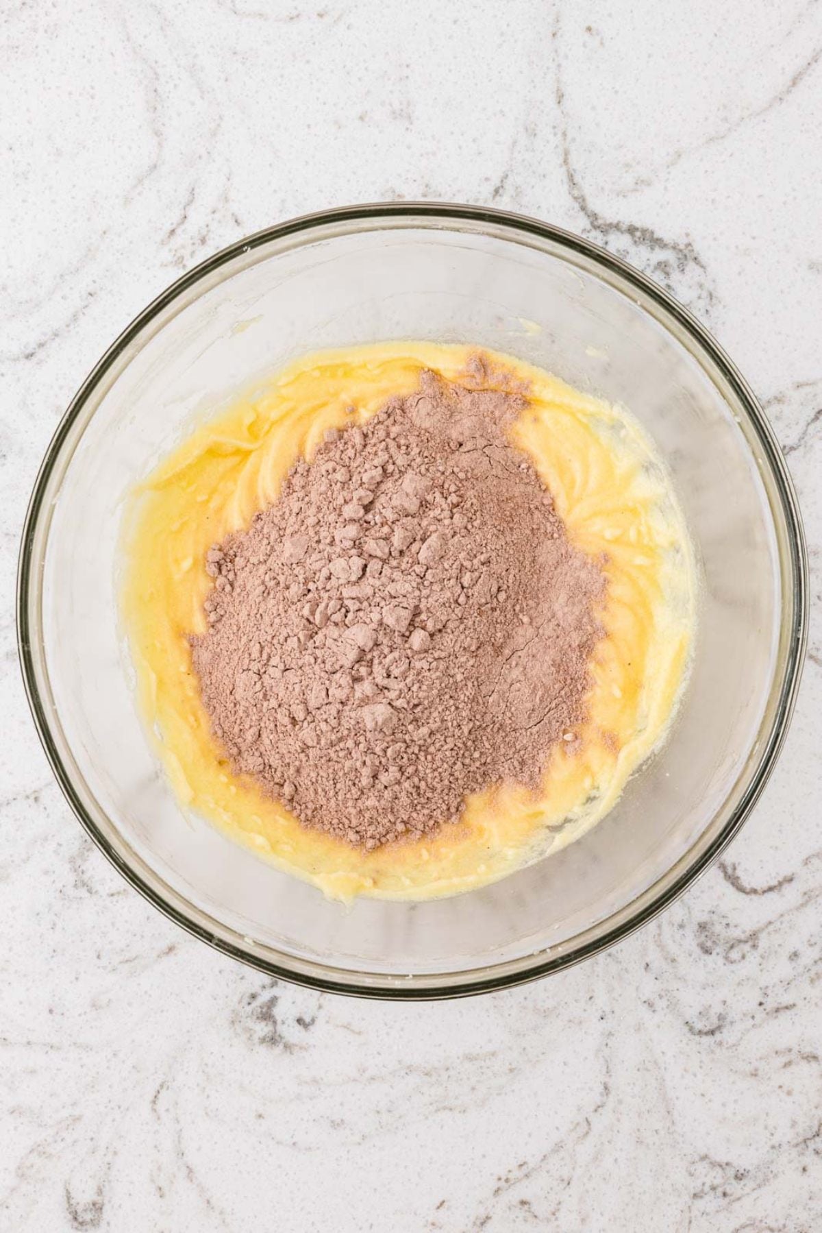 A glass bowl with yellow batter and a mound of cocoa powder on a white marbled surface.