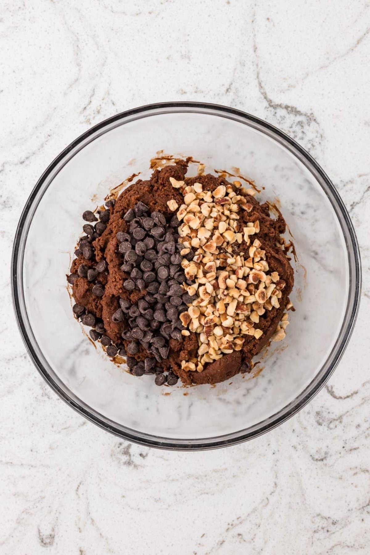 Glass bowl with cookie dough, chocolate chips, and chopped nuts on a white marble surface.