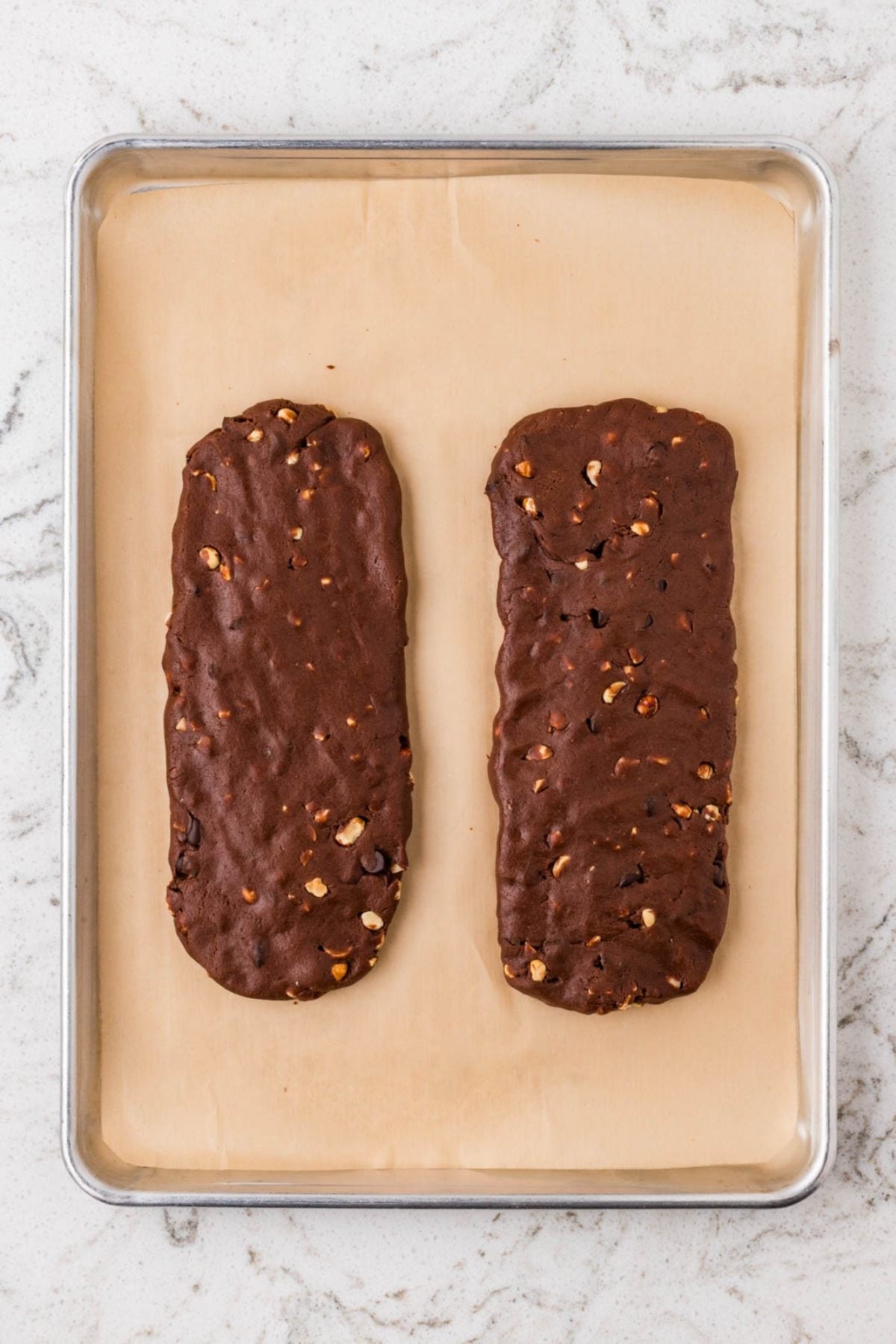 Two unbaked chocolate biscotti logs with nuts on a parchment-lined baking sheet.