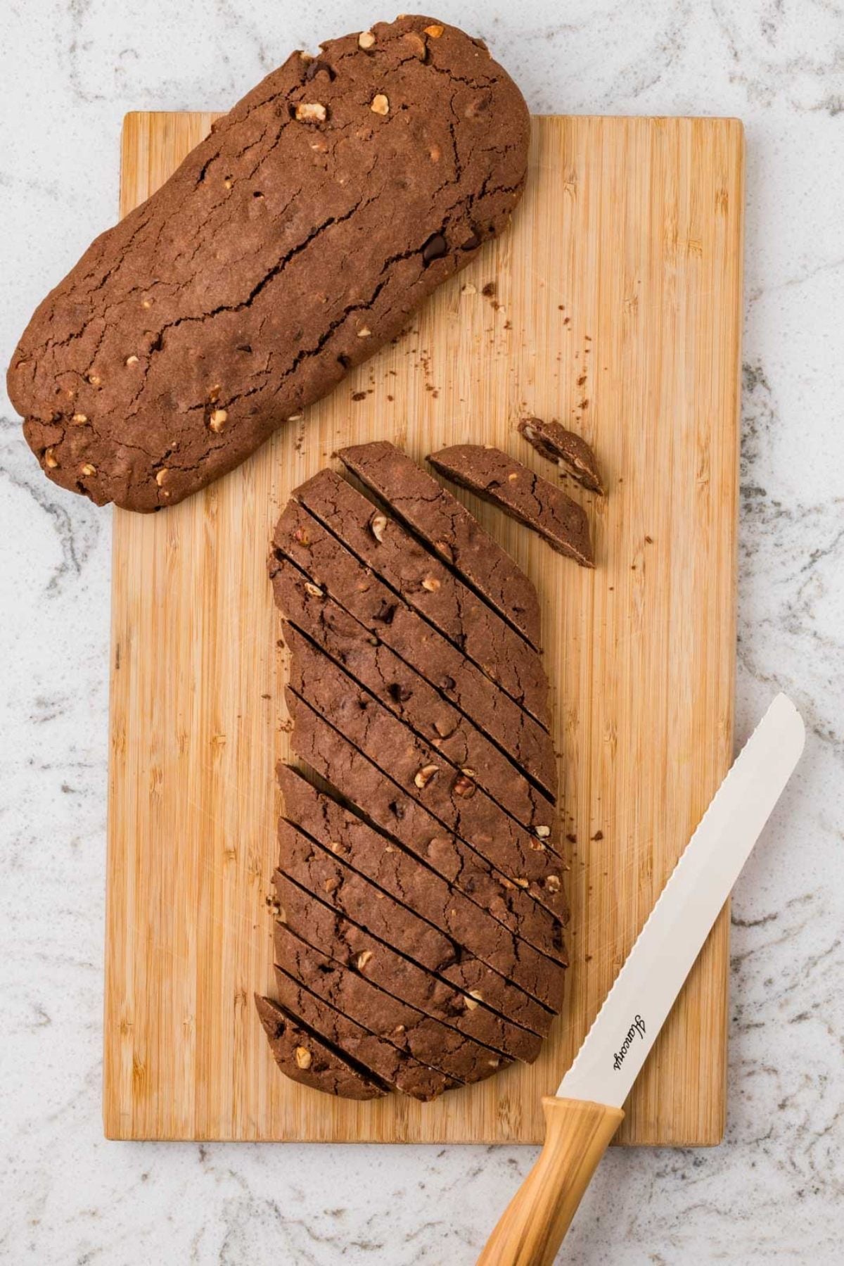 Two chocolate biscotti loaves, one sliced, on a wooden cutting board with a serrated knife beside them.